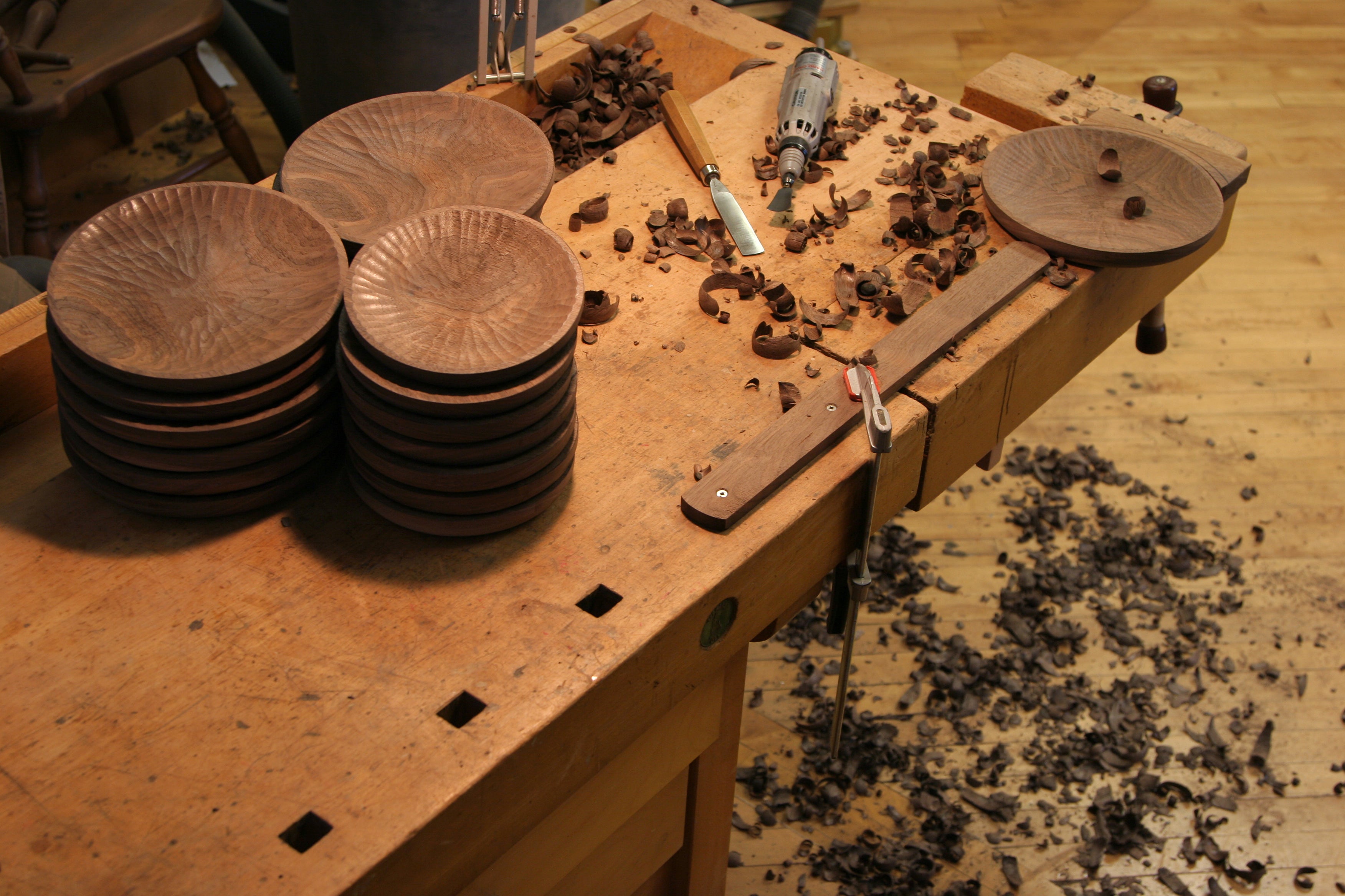 Woodworking bench with wooden plates, tools, and wood shavings on a wooden floor.