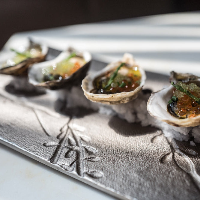Five raw oysters topped with orange roe, green herbs, and white caviar, served on a textured silver platter resting on a bed of crushed ice.