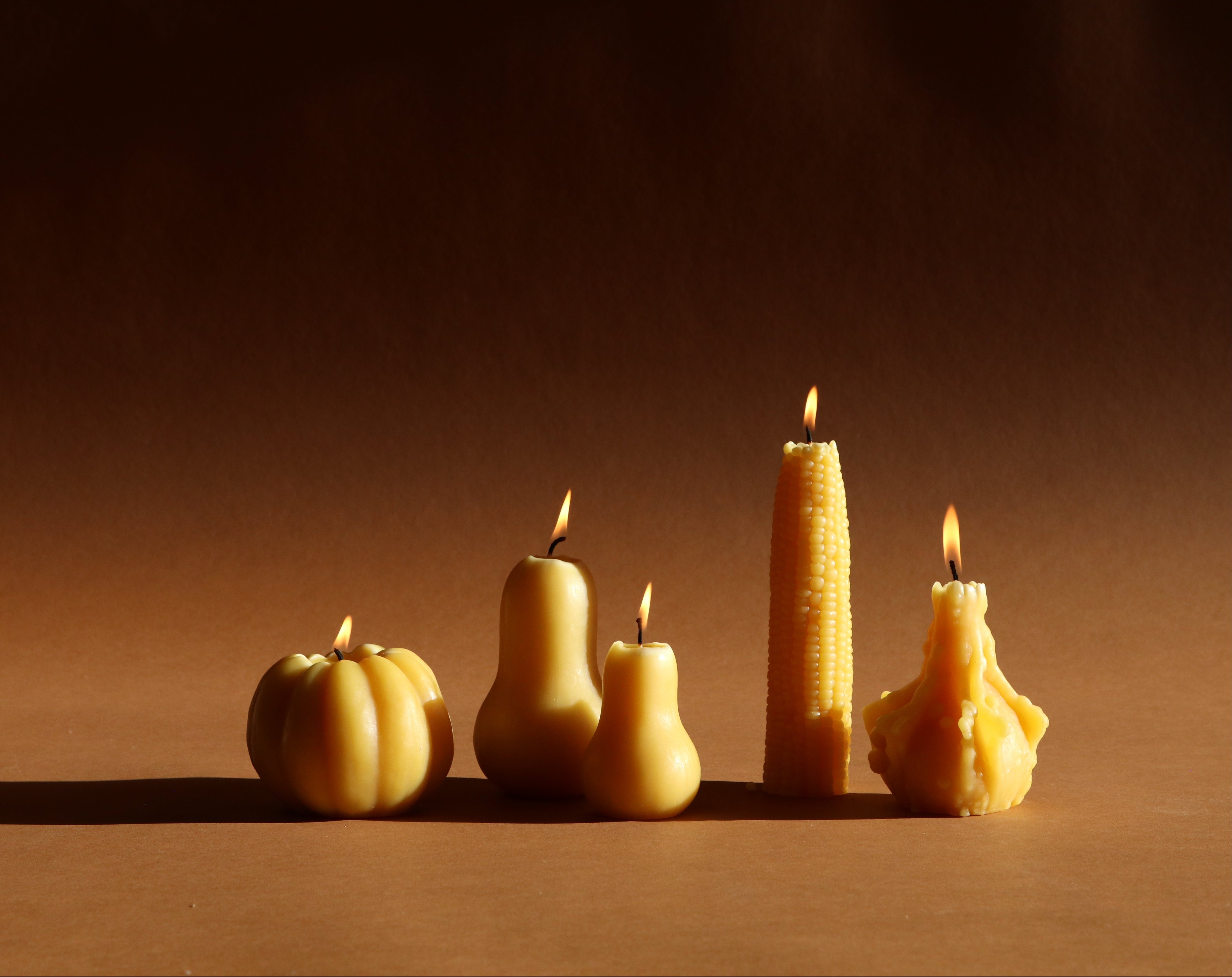 Four novelty beeswax candles shaped like a pumpkin, pear, corn cob, and a raw bee, lit and standing on a brown surface against a dark brown background.