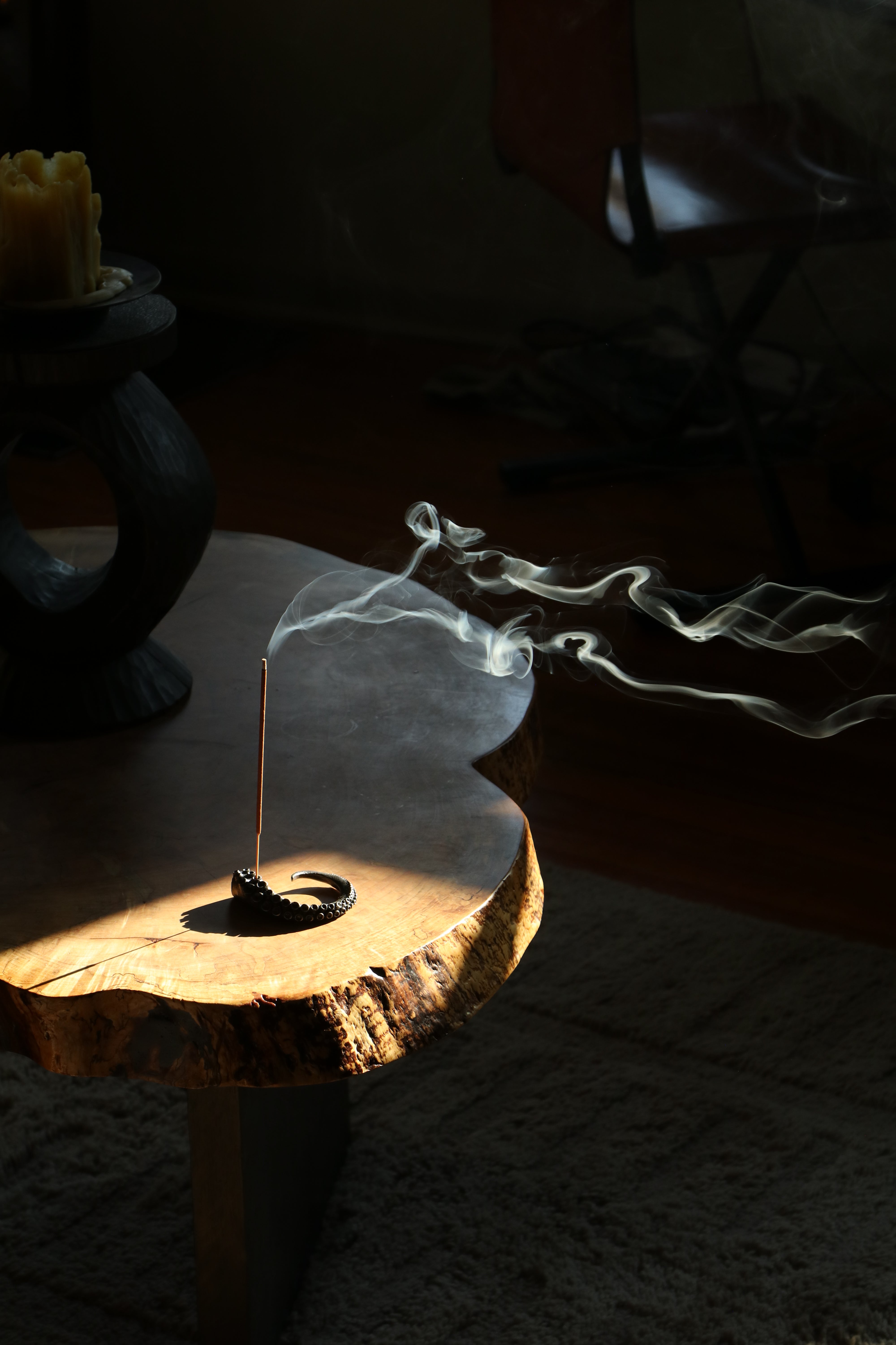 A single incense stick burns in a small black incense holder set on a rustic live-edge wooden table. Wispy white smoke curls through the air, and the scene is lit by dramatic, low-angle sunlight in a dim interior with a blurred chair in the background.