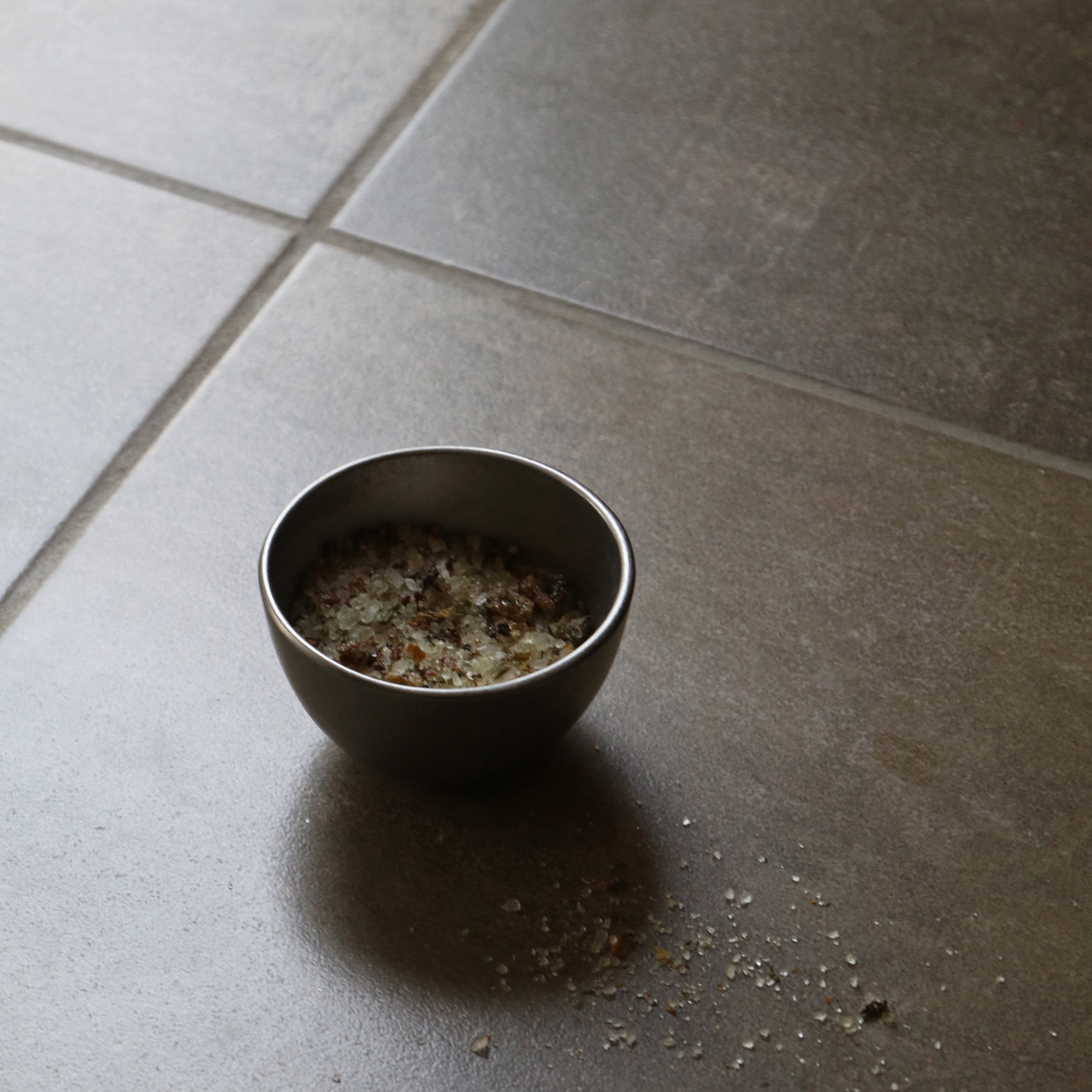 A small dark metal bowl filled with coarse salt or seasoning, placed on a dark gray tiled floor with some spilled seasoning scattered nearby.