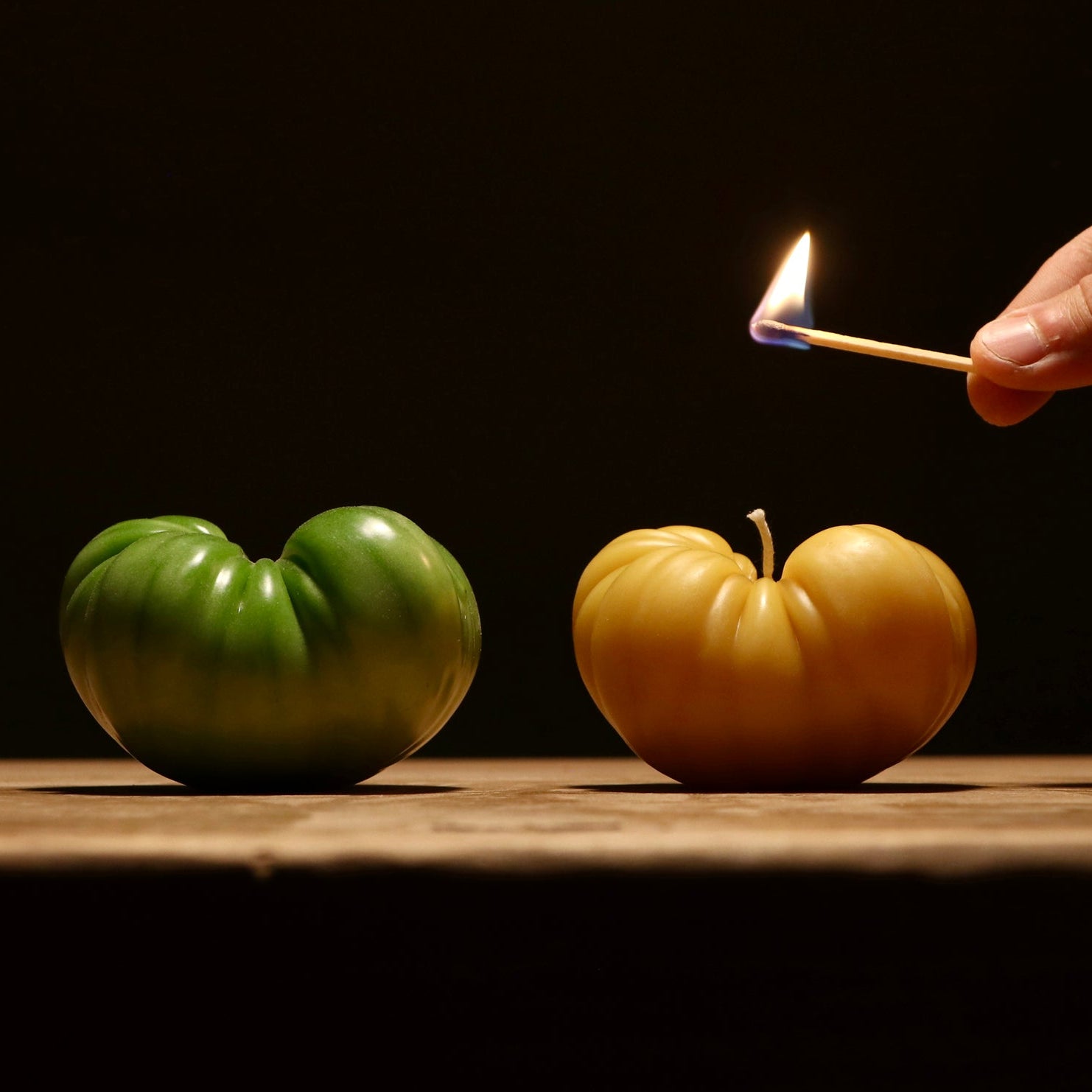 Two green and yellow tomatoes on a wooden surface with a hand holding a match.