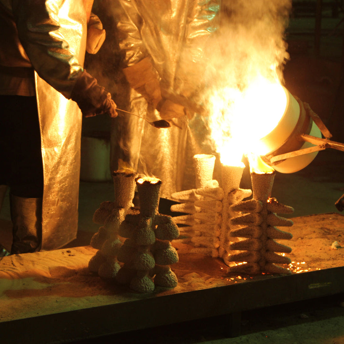 Workers in protective suits handle a glowing molten metal crucible over stacked ceramic molds on a worktable in a dark industrial setting.