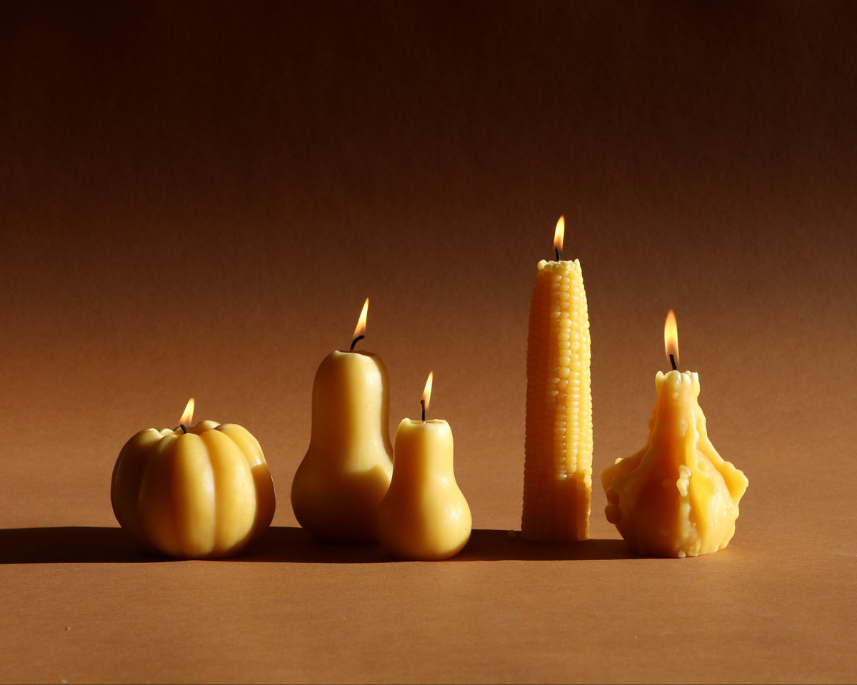 Four novelty beeswax candles shaped like a pumpkin, pear, corn cob, and a raw bee, lit and standing on a brown surface against a dark brown background.