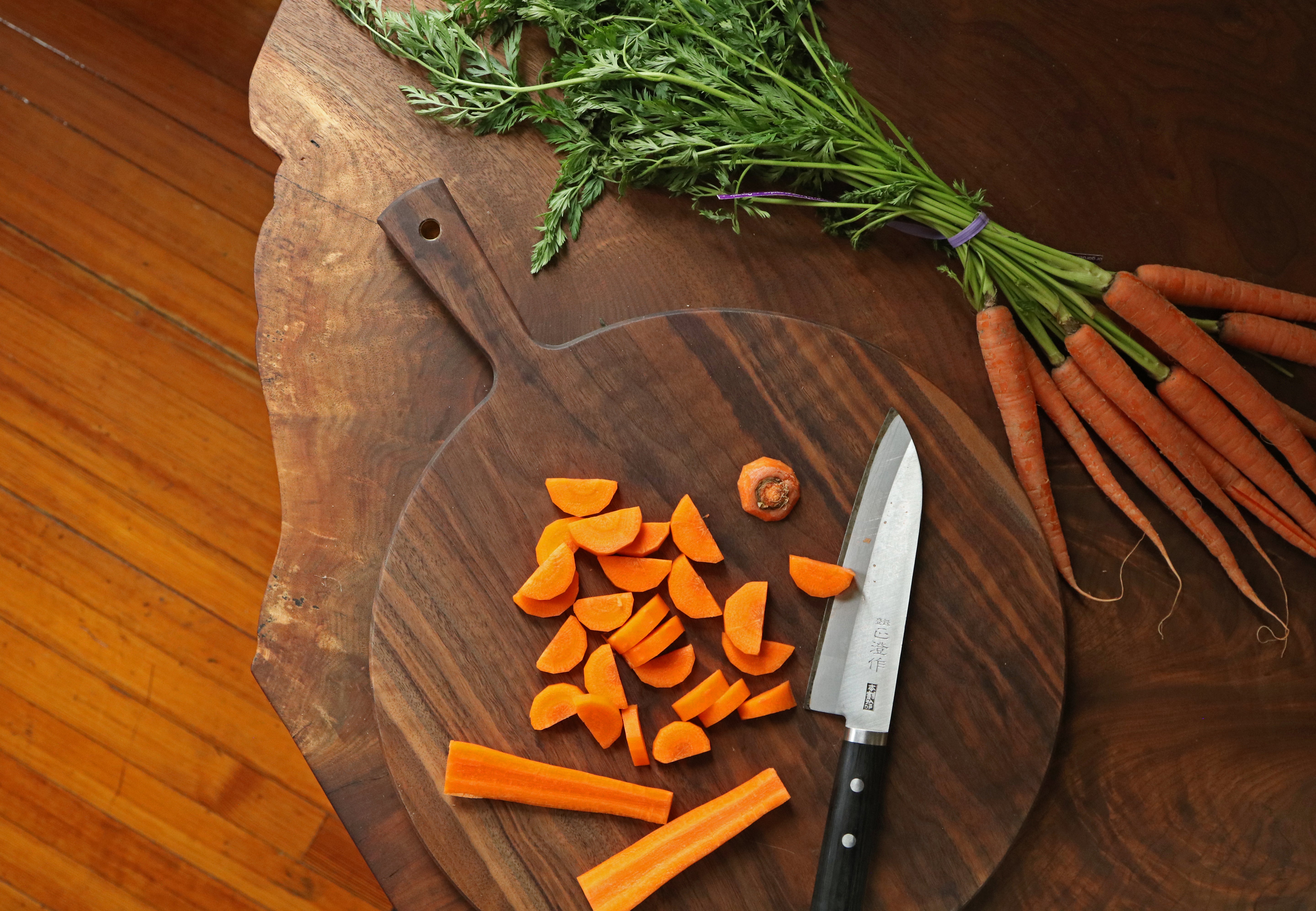Sliced carrots on a wooden cutting board with a knife and whole carrots.
