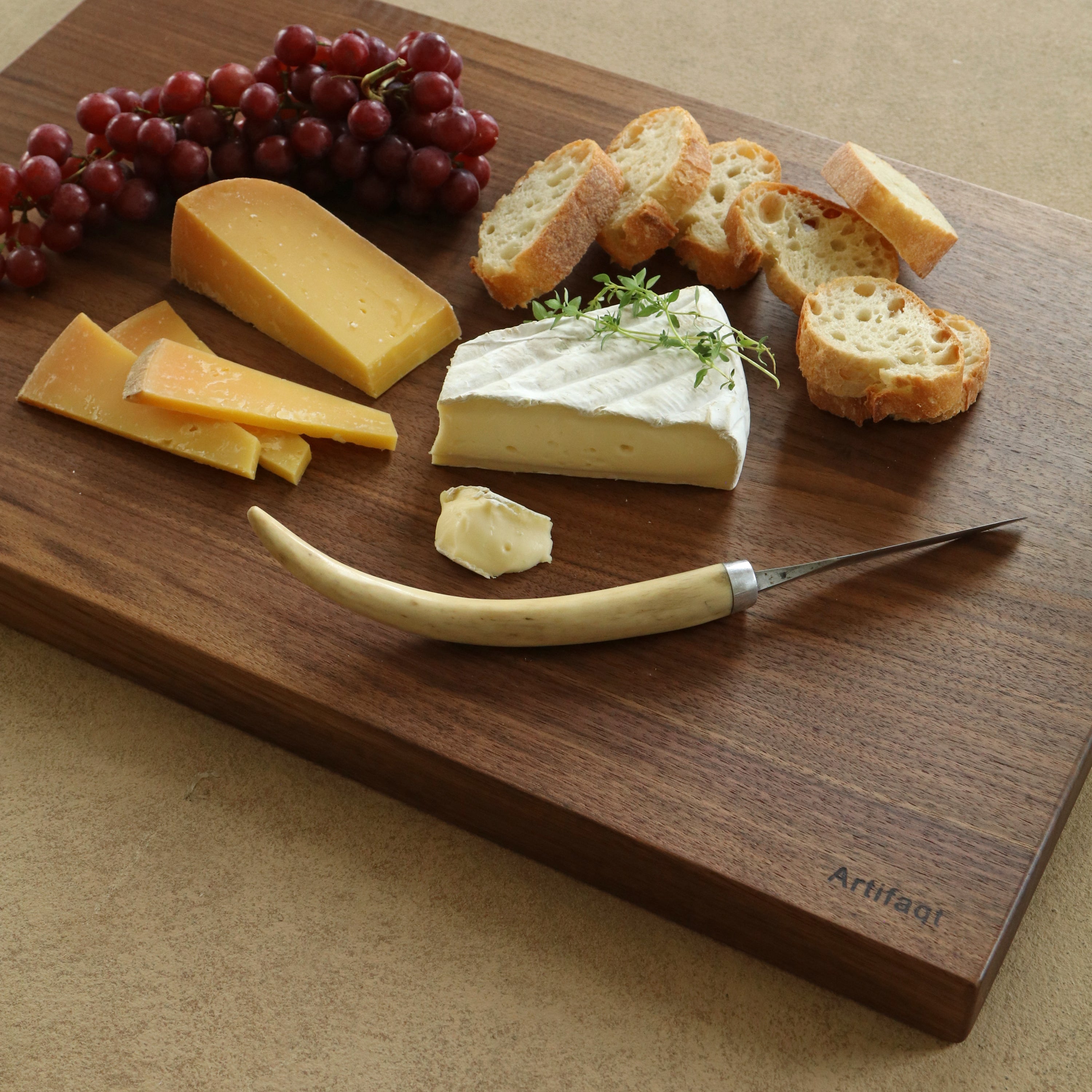 Wooden cutting board with cheese, bread, and grapes on a beige surface