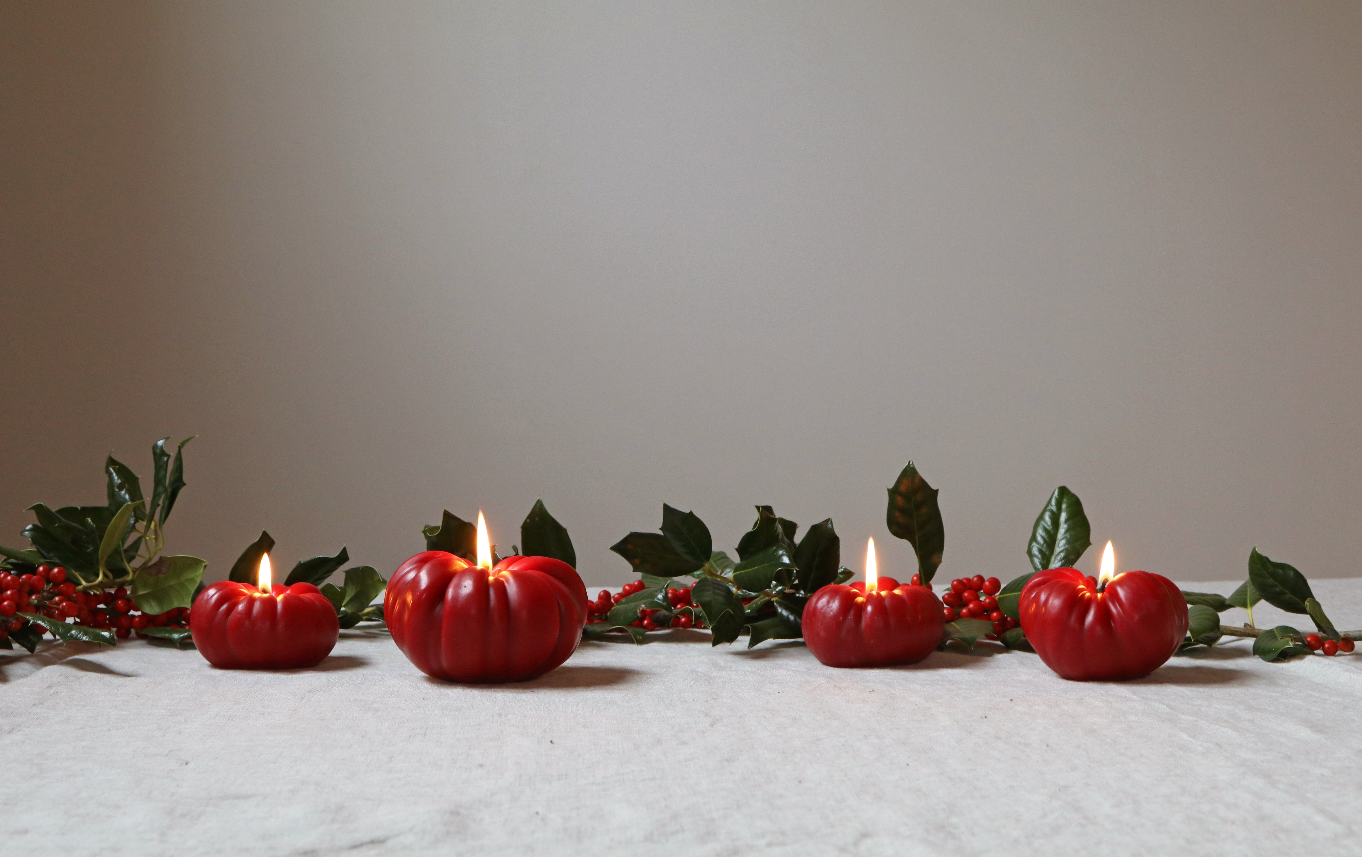 Red tomato-shaped candles with green leaves and berries on a white surface.