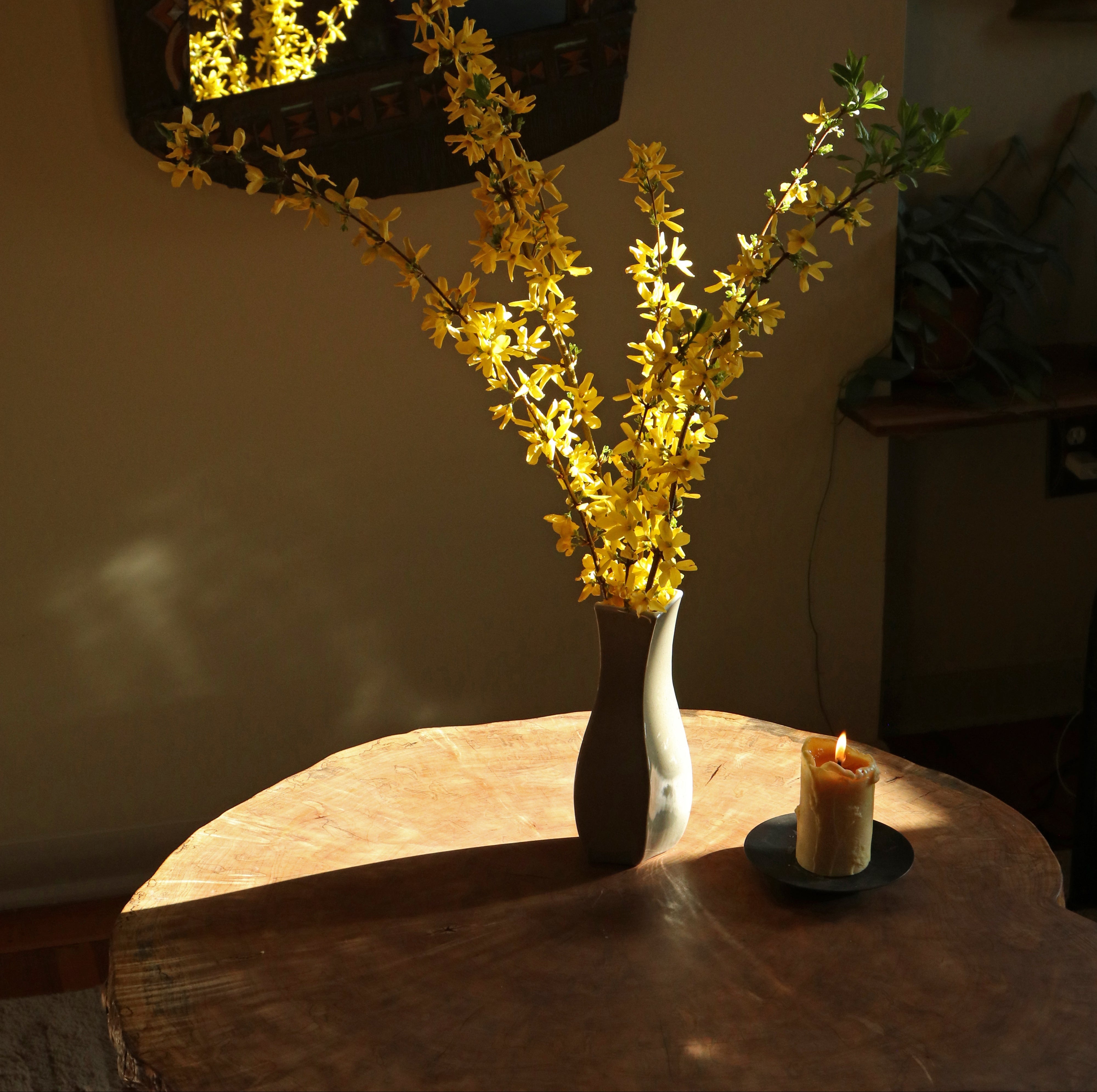Vase with yellow flowers on a wooden table with a candle, against a neutral wall.