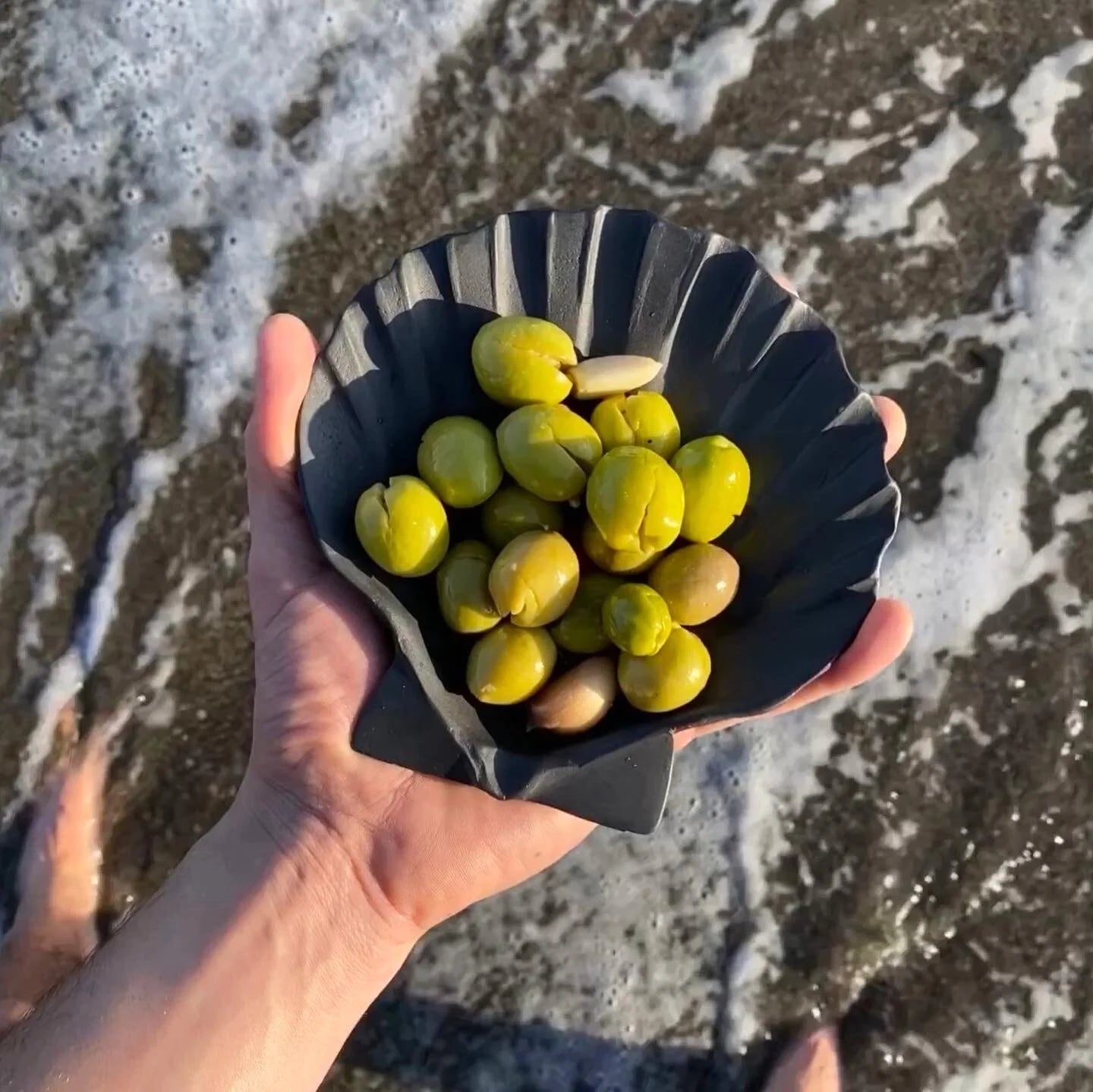 Hand holding a black bowl with green fruits against a beach background