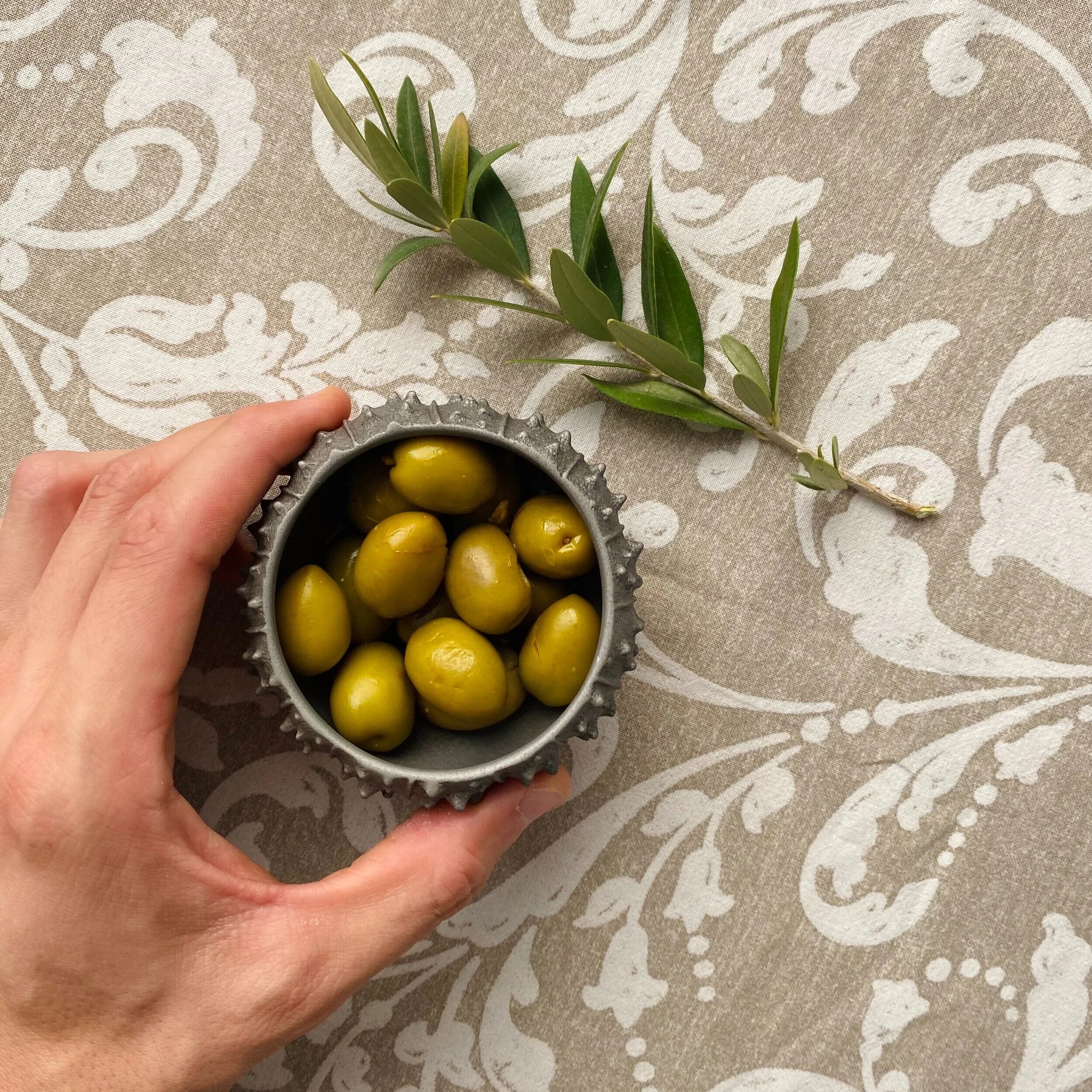 Hand holding a small metal container of green olives with an olive branch on a patterned background